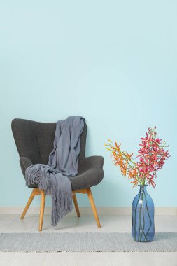 Rahat battaniyeli gri koltuk ve mavi duvarın yanındaki vazoda çiçekler.