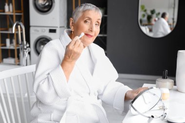 Yaşlı kadın banyoda makyaj yapıyor.