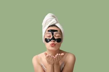 Yüzünde aktif kömür maskesi olan genç bir kadın yeşil arka plana öpücük yolluyor.