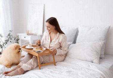 Young woman with cute Pomeranian dog having breakfast in bedroom