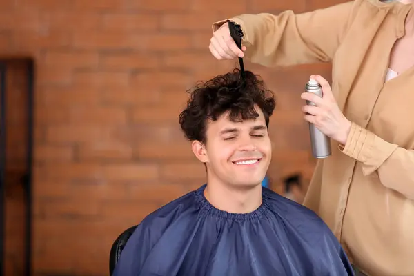 Professional hairdresser applying hair spray on young man\'s curly hair in beauty salon