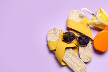 Parmak arası terlikli güneş gözlüğü, mini şemsiye ve leylak arka planda bir şişe güneş kremi.