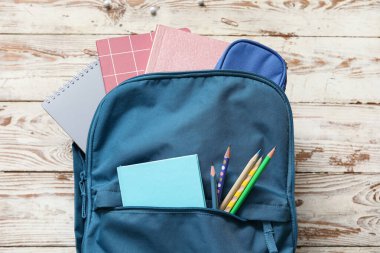 Mavi okul çantası, üzerinde defterler ve tahta arka planda kalemler var.