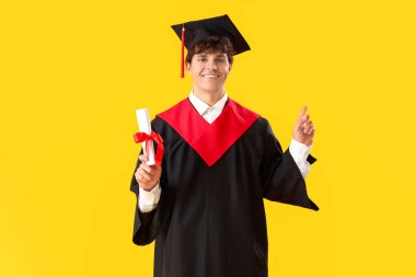 Sarı arka plandaki bir şeyi işaret eden diplomalı bir erkek öğrenci.
