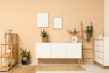 Odada bej duvarın yanında merdiven ve ev bitkileri olan modern beyaz çekmeceler.