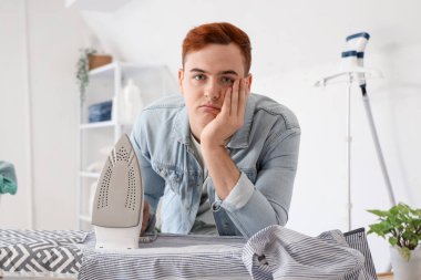 Çamaşır odasında ütü tahtasına yaslanmış elektrikli ütüyle sıkılmış genç bir adam