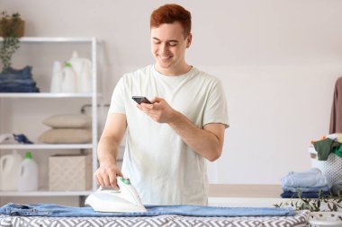 Cep telefonlu yakışıklı genç adam çamaşır odasında giysileri ütülüyor, yakın plan.