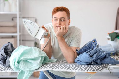 Çamaşır odasında ütü masasına yaslanmış elektrikli ütü ve buruşuk giysilerle sıkılmış genç bir adam.