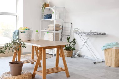 Çamaşır odasının içinde ütü masası, sepet içinde giysiler ve saksı var.