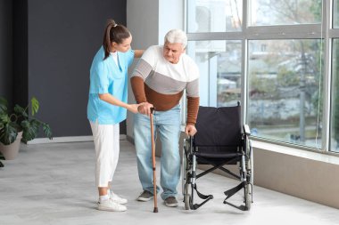 Hemşire yaşlı adama hastanede tekerlekli sandalyeye oturması için yardım ediyor.