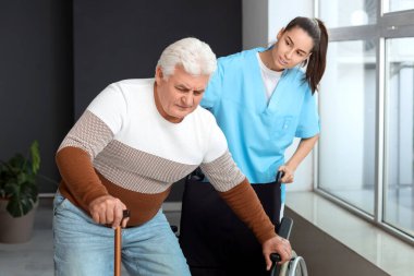 Hemşire yaşlı adama hastanede tekerlekli sandalyeye oturması için yardım ediyor.