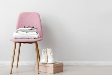 Katlanmış süveter yığını. Pembe sandalyede kitap ve beyaz duvarın yanında botlar.