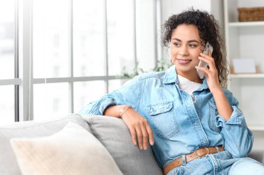 Young African-American woman talking by mobile phone on sofa at home