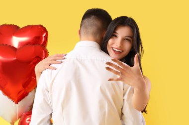 Alyansı ve kalp şeklinde balonları olan nişanlı genç çift sarı arka planda sarılıyorlar. Sevgililer Günü kutlaması