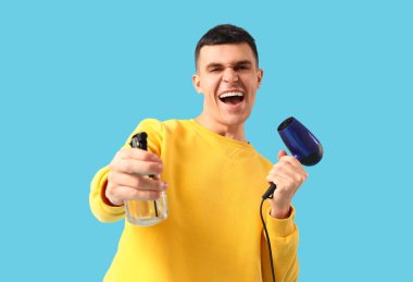 Saç kurutma makinesi ve mavi arka planda sprey şişesi olan yakışıklı bir genç.