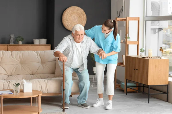 Nurse helping senior man with stick to stand up from sofa at home