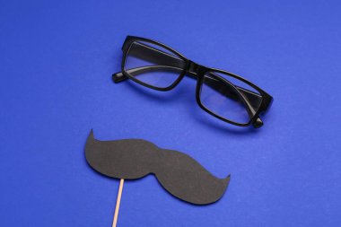 Kağıt bıyıklı tahta sopa ve mavi arka planda gözlük.