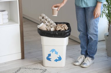 Mutfakta geri dönüşüm logosuyla dolu bir çöp kutusuna kağıt bardak atan kadın.