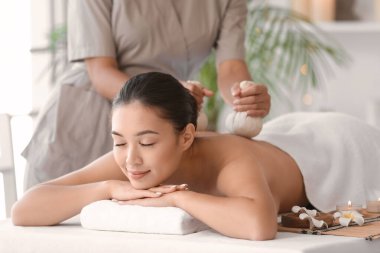 Genç Asyalı kadın spa salonunda bitki torbalarıyla masaj yaptırıyor.