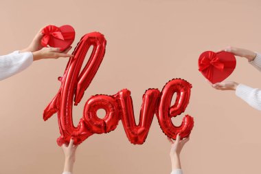 Kalp şeklinde hediye kutularını tutan kadın eller ve bej arka plandaki balonlardan yapılmış aşk kelimesi. Sevgililer Günü kutlaması