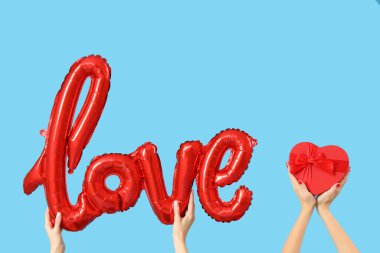 Kalp şeklinde hediye kutusu tutan kadın elleri ve mavi arka plandaki balonlardan yapılmış aşk kelimesi. Sevgililer Günü kutlaması