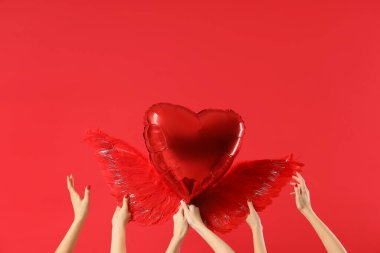 Kırmızı arka planda Aşk Tanrısı 'nın kanatları olan kalp şeklinde balon tutan kadın eller. Sevgililer Günü kutlaması
