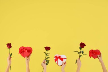 Kalp şeklinde hediye kutuları ve sarı arka planda güller tutan kadın elleri. Sevgililer Günü kutlaması