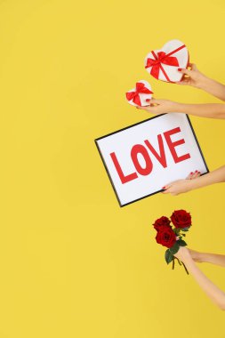 Kadın elleri çerçeveyi tutarken sevgi dolu, kalp şeklinde hediye kutuları ve sarı arka planda güller. Sevgililer Günü kutlaması