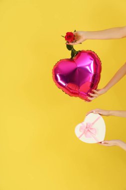 Kalp şeklinde balon, hediye kutusu ve sarı arka planda gül tutan kadın elleri. Sevgililer Günü kutlaması