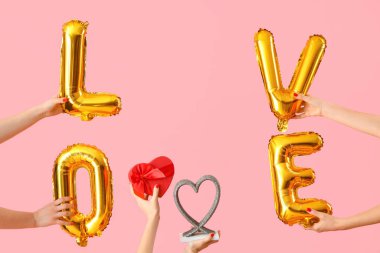 Balonlardan, hediye kutusundan ve pembe arka planda dekoratif kalpten yapılmış aşk sözcüğünü tutan kadın eller. Sevgililer Günü kutlaması