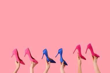 Pembe arka planda farklı yüksek topuklu ayakkabılar tutan kadın eller.