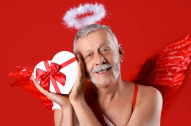 Aşk tanrısı gibi giyinmiş kırmızı arka planda hediye kutusu olan olgun bir adam, yakın plan. Sevgililer Günü kutlaması