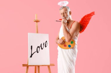 Aşk tanrısı gibi giyinmiş düşünceli olgun bir adam elinde boya ve pembe arka planda aşk sözcüğüyle sehpa tutuyor. Sevgililer Günü kutlaması