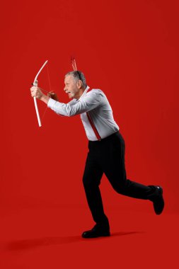 Aşk tanrısı gibi giyinmiş olgun bir adam kırmızı arka planda ok atıyor. Sevgililer Günü kutlaması