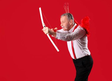 Aşk tanrısı gibi giyinmiş olgun bir adam kırmızı arka planda ok atıyor. Sevgililer Günü kutlaması