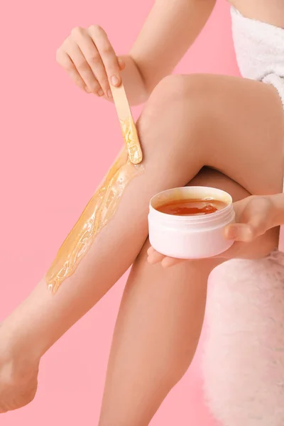 Young woman applying sugaring paste onto her legs against pink background