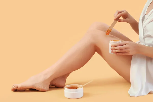 Young woman applying sugaring paste onto her legs against beige background