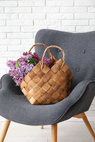 Basket with beautiful lilac flowers on armchair near light brick wall, closeup