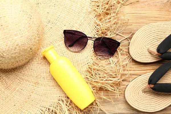 Terlikli güneş gözlüğü, hasır şapka ve ahşap arka planda bir şişe güneş kremi.