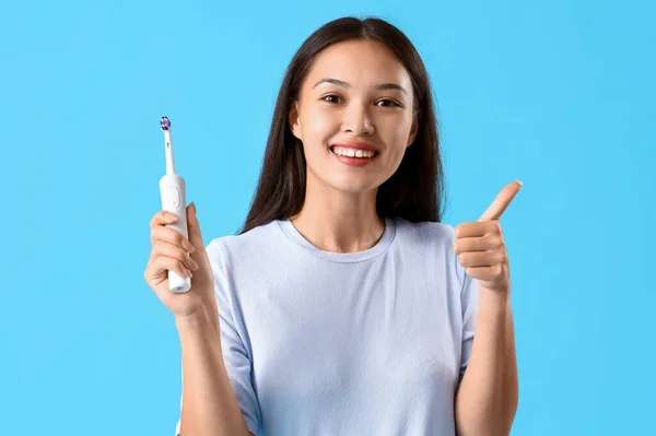 青い背景に親指を示す電気歯ブラシを持つ若いアジアの女性を笑う — ストック写真