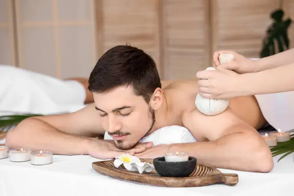 Young man getting massage with herbal bags in spa salon