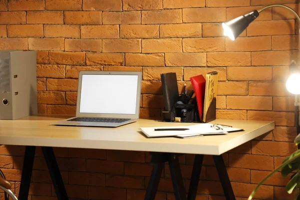Blank laptop on table and glowing lamp in office at night