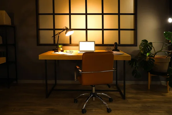 Interior of dark office with workplace, blank laptop and glowing lamp at night