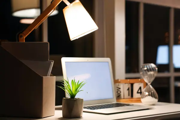 Glowing lamp with folder and plant on desk in office at night, closeup