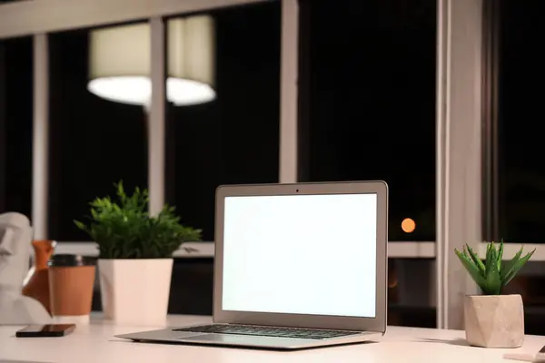 Blank laptop and plants on desk in office at night, closeup