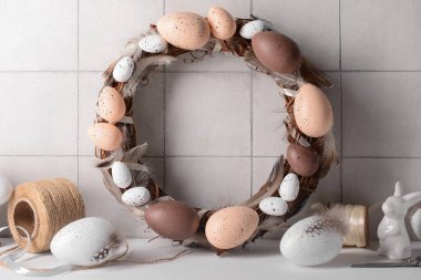 Yumurta ve tüylerden yapılmış Paskalya çelengi. Beyaz duvarın yanındaki masada erzak var.