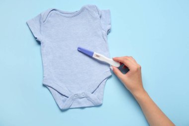 Pozitif hamilelik testi ve bebek tulumu olan bir kadın eli.