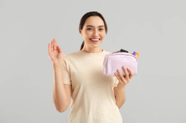 Gri arka planda el sallayan makyaj ürünleriyle dolu kozmetik çantasıyla güzel genç bir kadın.