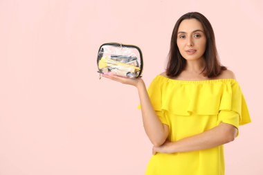 Pembe arka planda makyaj malzemeleriyle dolu kozmetik çantası olan güzel genç bir kadın.