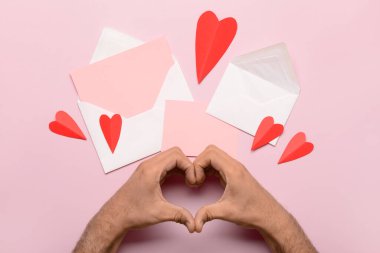 Erkek eli, zarf ile kalp şekli, boş kart ve pembe arka planda kırmızı kağıt kalp yapıyor. Sevgililer Günü kutlaması
